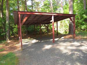 A wooden carport stands on a gravel surface in a wooded area with trees and sunlight filtering through the leaves. The carport is open on all sides with a metal roof supported by wooden beams.