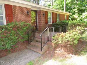 A brick house with red shutters and a black front door surrounded by green bushes. A small set of brick steps with black railings leads to the entrance. The area is shaded by trees.