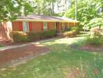 Located at 405 S Elm Street in Creedmoor, this brick house with red shutters is surrounded by lush greenery and trees. It features a quaint front porch with steps and a metal railing, bordered by bushes and a grassy lawn adorned with flower beds. The setting is sunny and tranquil.