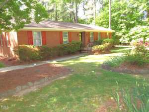 Located at 405 S Elm Street in Creedmoor, this brick house with red shutters is surrounded by lush greenery and trees. It features a quaint front porch with steps and a metal railing, bordered by bushes and a grassy lawn adorned with flower beds. The setting is sunny and tranquil.