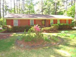 A single-story brick house with red shutters is surrounded by a lush green yard and tall trees. A small garden with blooming pink flowers is in the foreground, and the scene is illuminated by bright sunlight.