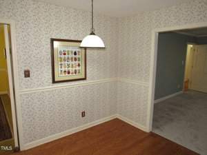 A small room features floral wallpaper, a framed vegetable poster, and a hanging light fixture. The floor is wooden, and an open doorway leads to a carpeted room with a closed door visible.