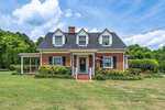 A charming red-brick house with white trim and a steep, dark gray roof sits at 216 Grove Street in Oxford. It features dormer windows, a white porch, and a well-maintained lawn and garden. A flag hangs near the entrance, surrounded by trees under a partly cloudy sky.