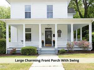 A white two-story house with a large, charming front porch. The porch features a swing, two potted plants, and white railing. Lush greenery surrounds the property, enhancing its inviting appearance.