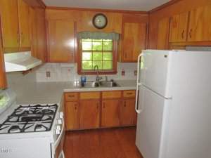 A small kitchen with wooden cabinets, a gas stove, a white refrigerator, and a double sink under a window. A clock hangs above the window, and the countertop has a white tile backsplash. The floor is wooden.