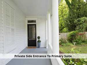 A white porch with columns and a black door, featuring a potted plant beside the entrance. The porch leads to a garden with a wooden fence and greenery. The text reads, "Private Side Entrance To Primary Suite.