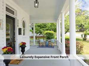 A spacious front porch with white pillars and a blue ceiling, featuring a cozy seating area with striped chairs and a small table. Potted flowers add color, and lush greenery surrounds the area. A mailbox and house number are visible.