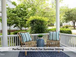A cozy front porch with two wicker chairs featuring blue and white striped cushions. A small blue side table with a green potted plant sits in between. Lush greenery surrounds the porch, creating a peaceful, summer ambiance.