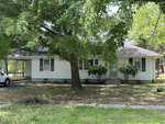 A small, single-story house located at 306 W C Street in Butner features white siding and green shutters, surrounded by trees and lush grass. A large tree shades the front yard, with a driveway leading to a carport on the left side of the home.