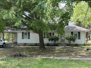 A small, single-story house located at 306 W C Street in Butner features white siding and green shutters, surrounded by trees and lush grass. A large tree shades the front yard, with a driveway leading to a carport on the left side of the home.
