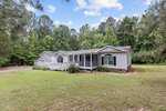 A single-story house at 4161 Cannady Mill Road, Oxford, boasts a light gray exterior and a gabled roof, nestled among lush green trees. The home features a charming front porch and a well-maintained lawn. A gravel driveway invites you to the property under a partly cloudy sky.