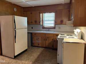 A small kitchen with wooden cabinets, a white refrigerator, an electric stove, and a sink under a window with blinds. The floor is covered with vinyl tiles, and the walls have wood paneling.