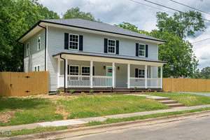 Nestled at 110 1st Street, Oxford, this two-story white house with a dark roof boasts a wide front porch and black shutters. Encircled by a wooden fence, it sits among lush grass and trees. A charming pathway leads to the front door, while a sidewalk lines the street in front.