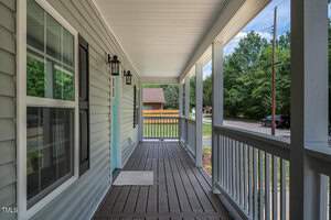 A wooden front porch with a railing and a covered roof. The porch features a doormat and two wall-mounted lanterns. There's a view of the street lined with trees and a parked car. A house with a sloped roof is visible in the background.