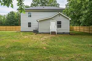 Back view of a two-story house with gray siding, white trim, and a small wooden staircase leading to a white door. The house is surrounded by a wooden fence and green lawn, with trees in the background.
