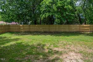 A lush green backyard with a wooden fence enclosing the space. Dense trees line the background, providing shade and privacy. The grass is partially sunlit, showing patches of sunlight and shade.