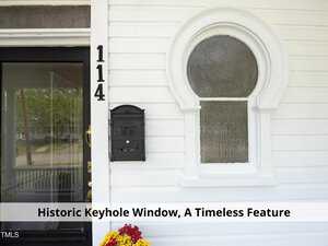 A white house exterior featuring a black mailbox, the number "114," and a distinctive keyhole-shaped window. Below the window is a planter with small yellow and red flowers. The image includes the caption, "Historic Keyhole Window, A Timeless Feature.