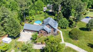 Aerial view of a large brick house surrounded by tall trees and greenery. The property features a circular above-ground pool in the backyard and a long driveway leading to a spacious garage. Neighboring homes are visible in the lush landscape.