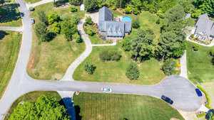 Aerial view of a suburban neighborhood featuring a large house with a swimming pool. The house is surrounded by trees and a well-maintained lawn. A few cars are parked on the street, and a curved driveway leads to the house.