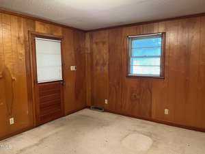 An empty room with wood-paneled walls, a single wooden door to the left with a window above it, and another window to the right. Beige carpet covers the floor. The room appears well-lit by natural light from the windows.