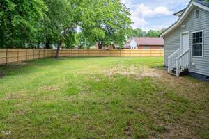 A spacious backyard with a large lawn, surrounded by a wooden fence. There are a few trees providing shade. The back of a house with gray siding and white steps is seen on the right. The sky is partly cloudy, with patches of blue visible.