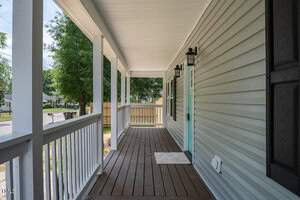 A cozy porch with a white ceiling and light gray siding. It features dark wooden flooring, a mint green door, and two black lantern-style wall lights. The porch overlooks a quiet street lined with trees.