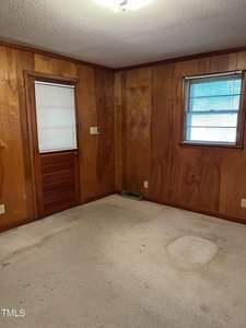 An empty room with wood-paneled walls and a light-colored carpet. There is a wooden door with a small window on the left and another window on the right, both with blinds. A ceiling light fixture is visible. The room appears to be in need of renovation.