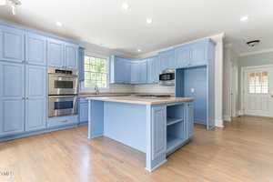 A spacious kitchen featuring light blue cabinetry, a central island with a stovetop, stainless steel double ovens, and a microwave. The room is brightened by natural light from a window above the sink. Hardwood floors and recessed lighting complete the look.