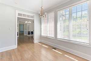 A bright, empty room with light hardwood floors and large windows with blinds, allowing natural light to flood in. The room is decorated with modern light fixtures and opens into another room with double doors.
