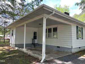 A small, single-story white house with a covered porch. The house has white siding, a green door, and one window on the visible side. It is surrounded by trees and grass, with a gravel area in front of the porch.