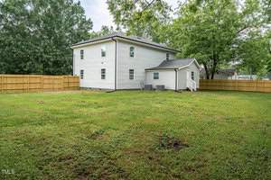 A two-story white house with a small attached structure at the back, surrounded by a wooden fence. The yard is spacious with green grass and a few trees, creating a serene outdoor space.