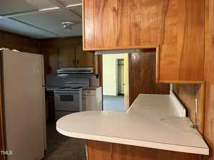 A small, rustic kitchen with wooden cabinets, a white refrigerator, and an electric stove. A counter with a rounded edge is in the foreground. The room has wood-paneled walls and a ceiling light fixture.