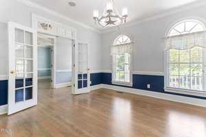 A bright, empty room featuring hardwood floors, white and blue walls, and arched windows with floral curtains. A chandelier hangs from the ceiling. French doors open to another room with similar decor.