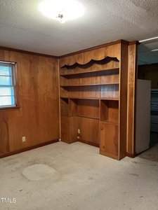 A room with wood-paneled walls and beige carpet features a built-in wooden shelving unit with three large shelves and two cabinets. A small window with blinds is on the left wall, and a ceiling light fixture is above.