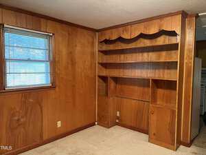 A room with wood-paneled walls featuring built-in shelving and cabinets. A window with blinds is on the left, and beige carpeting covers the floor. Electrical outlets are visible on the walls.