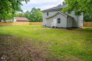 A two-story house with gray siding and a shingled roof, surrounded by a spacious grassy backyard and wooden fence. There are trees bordering the property and a neighboring house visible in the distance. Clear skies overhead.