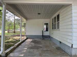 A covered porch with a concrete floor attached to a white house. There's a door with a window and several other windows on the right. A wooden fence and trees are visible in the background.