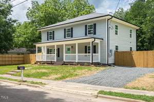 A two-story, white house with black shutters and a covered front porch. The gravel driveway leads to a fenced backyard. The yard is landscaped with young shrubs, and the mailbox is visible near the street. Trees are in the background.