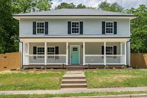 Two-story house with light gray siding and a green front door. Features a full-length front porch with white railings. The roof is dark gray, and the yard has a small pathway leading to the porch. Surrounded by a wooden fence and greenery.
