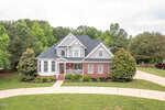 Located on 8803 Sunflower Meadows Lane in Wake Forest, this two-story suburban house with gray siding and brick accents features a gabled roof and white columns on the porch. It is surrounded by a lush green lawn, trees, and a curved driveway.