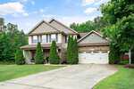 A two-story suburban house on 1174 Jackson Court boasts a brick and siding exterior, double garage, and driveway. The front yard features neatly clipped grass with tall trees near the entrance. Located in Creedmoor, the sky is partly cloudy with trees adorning the background.
