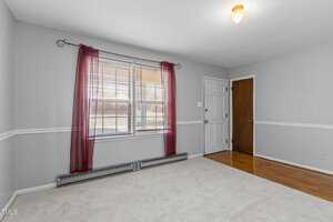 A small, empty room with gray walls and a wooden floor partially covered by a light-colored carpet. A window with red curtains and blinds lets in natural light. Two doors, one wooden and one white, are on opposite walls.