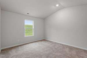 An empty room with beige carpet and light gray walls. A single window shows a grassy field outside. The ceiling is vaulted with a small recessed light.