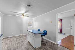 A minimalist room with gray walls and a wood-patterned floor. It features a small kitchen island with blue stools, a ceiling fan with lights, and a transition to a room with wooden flooring and red curtains.