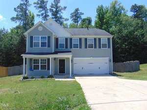 Located at 716 Pitch Pine Drive in Creedmoor, this two-story house boasts blue-gray siding and black shutters, complemented by a charming covered porch and a spacious double garage. The grassy front lawn and towering trees complete the picturesque setting under a clear blue sky.
