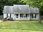 Located at 3700 Aztec Lane in Franklinton, this small, single-story gray house boasts white trim and a sloped roof. A charming white front door, dark shutters, and a small porch add character. A grassy yard with flower bushes and a tall tree is enclosed by a wooden fence surrounding the property.