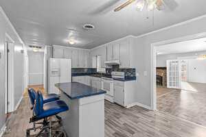 A kitchen with blue countertops and white cabinets. It includes a white stove, refrigerator, and a blue-tiled backsplash. Two blue bar stools sit at the counter. The room opens to a living area with wooden floors and a stone fireplace.
