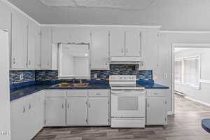 A kitchen with white cabinets, blue mosaic tile backsplash, and a blue countertop. It features a white electric stove with an oven and a double sink under a window. The flooring is wood-style laminate. An open doorway leads to another room.