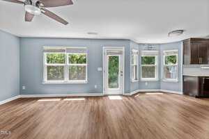 An empty room with light blue walls and wooden floors. It features three large windows, a glass door, and a ceiling fan. There is a partial view of a kitchen with dark cabinets to the right. Sunlight streams in, creating a bright atmosphere.