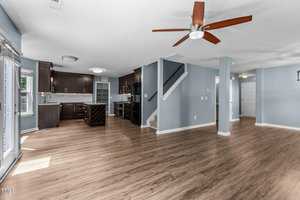 Spacious open-concept living area with light blue walls and wooden flooring. A kitchen with modern dark cabinets, marble countertops, and an island is on the left. A ceiling fan and stairs lead to the upper level. Natural light enters through large windows.